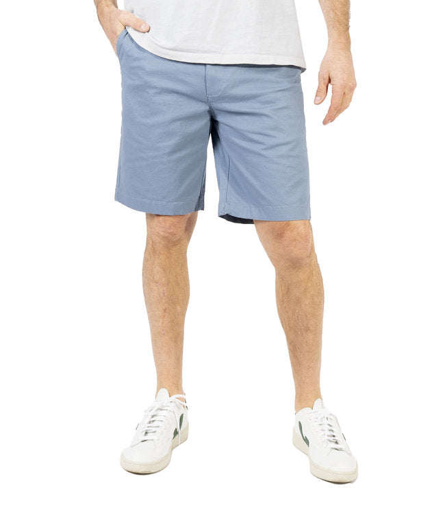 A person in a white shirt is sporting light blue Silver Jeans Ashton Stretch Twill Walking Shorts from Silver Jeans Co. and white sneakers with green accents. Their hands are in their pockets, and their head is out of frame. The background is plain white.