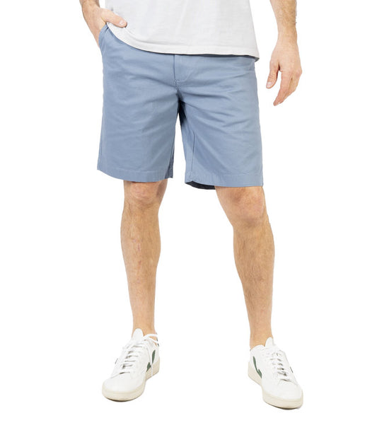 A person in a white shirt is sporting light blue Silver Jeans Ashton Stretch Twill Walking Shorts from Silver Jeans Co. and white sneakers with green accents. Their hands are in their pockets, and their head is out of frame. The background is plain white.