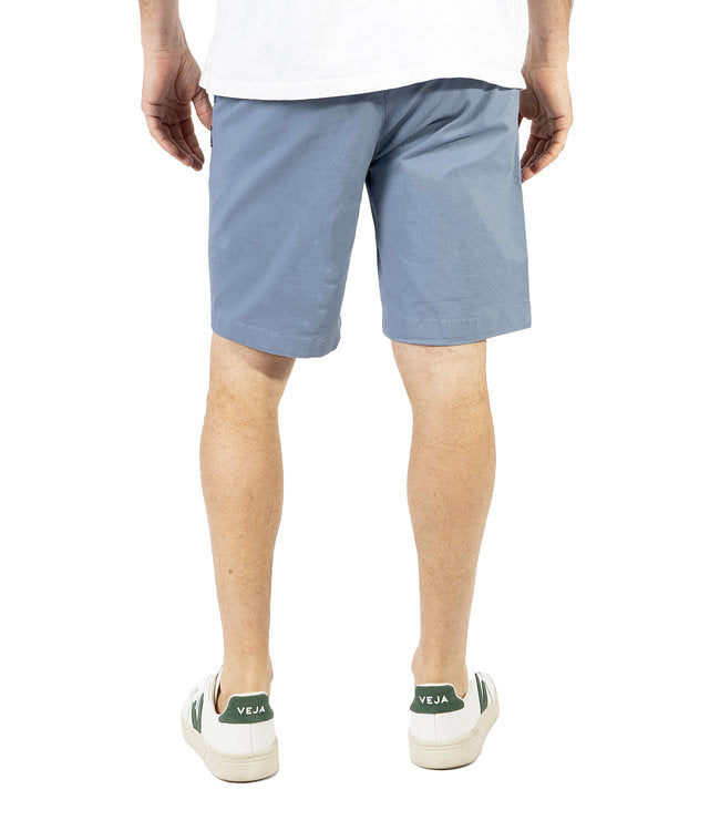 A person in a white shirt is sporting light blue Silver Jeans Ashton Stretch Twill Walking Shorts from Silver Jeans Co. and white sneakers with green accents. Their hands are in their pockets, and their head is out of frame. The background is plain white.