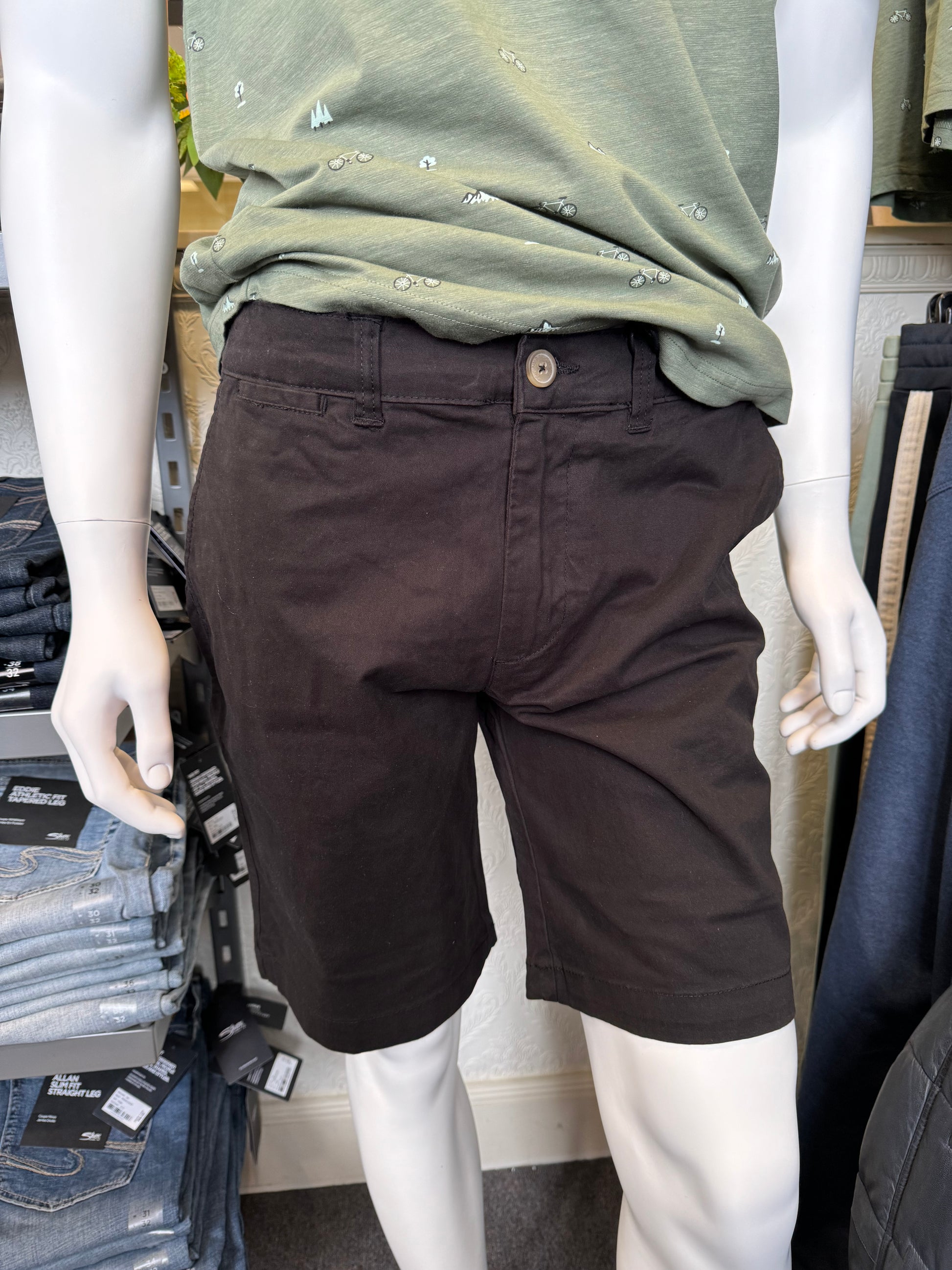 A mannequin dressed in a green shirt with a bicycle pattern and dark brown shorts is displayed in a store. In the background, Silver Jeans Ashton Stretch Twill Walking Shorts - Black and folded grey pants rest on a rack beside a quilted jacket. The store's textured wall is visible behind it all.