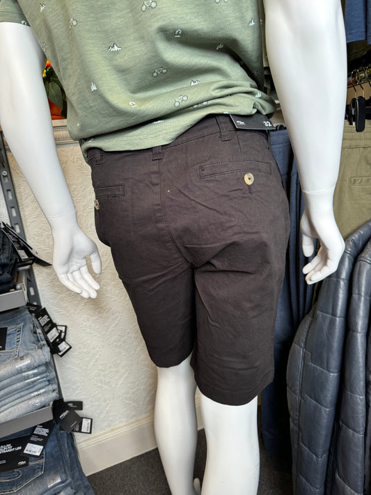 A mannequin dressed in a green shirt with a bicycle pattern and dark brown shorts is displayed in a store. In the background, Silver Jeans Ashton Stretch Twill Walking Shorts - Black and folded grey pants rest on a rack beside a quilted jacket. The store's textured wall is visible behind it all.