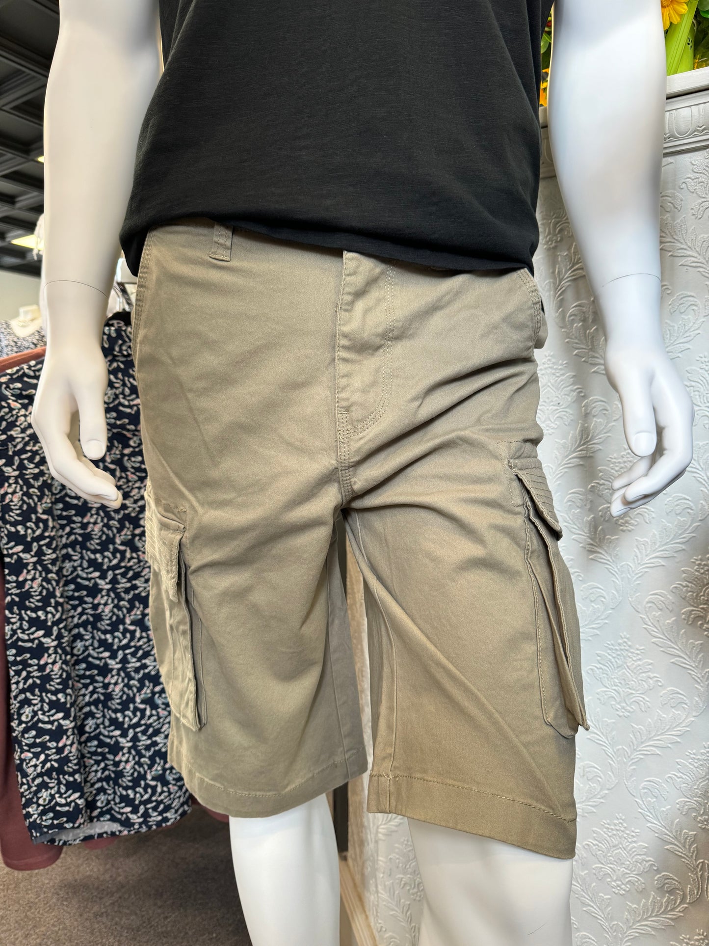 Close-up of the Silver Jeans Collin Classic Fit Cargo Bermudas in Light Olive, distinguished by a large side pocket, displayed on a mannequin. The fabric exhibits detailed stitching and the material looks exceptionally durable. In the background, shelves filled with folded clothes are visible.