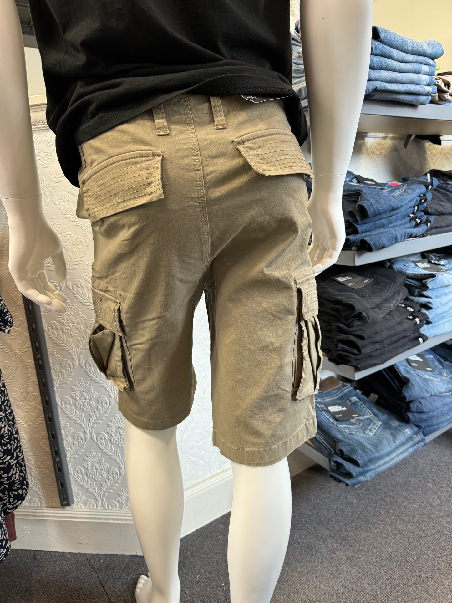 Close-up of the Silver Jeans Collin Classic Fit Cargo Bermudas in Light Olive, distinguished by a large side pocket, displayed on a mannequin. The fabric exhibits detailed stitching and the material looks exceptionally durable. In the background, shelves filled with folded clothes are visible.