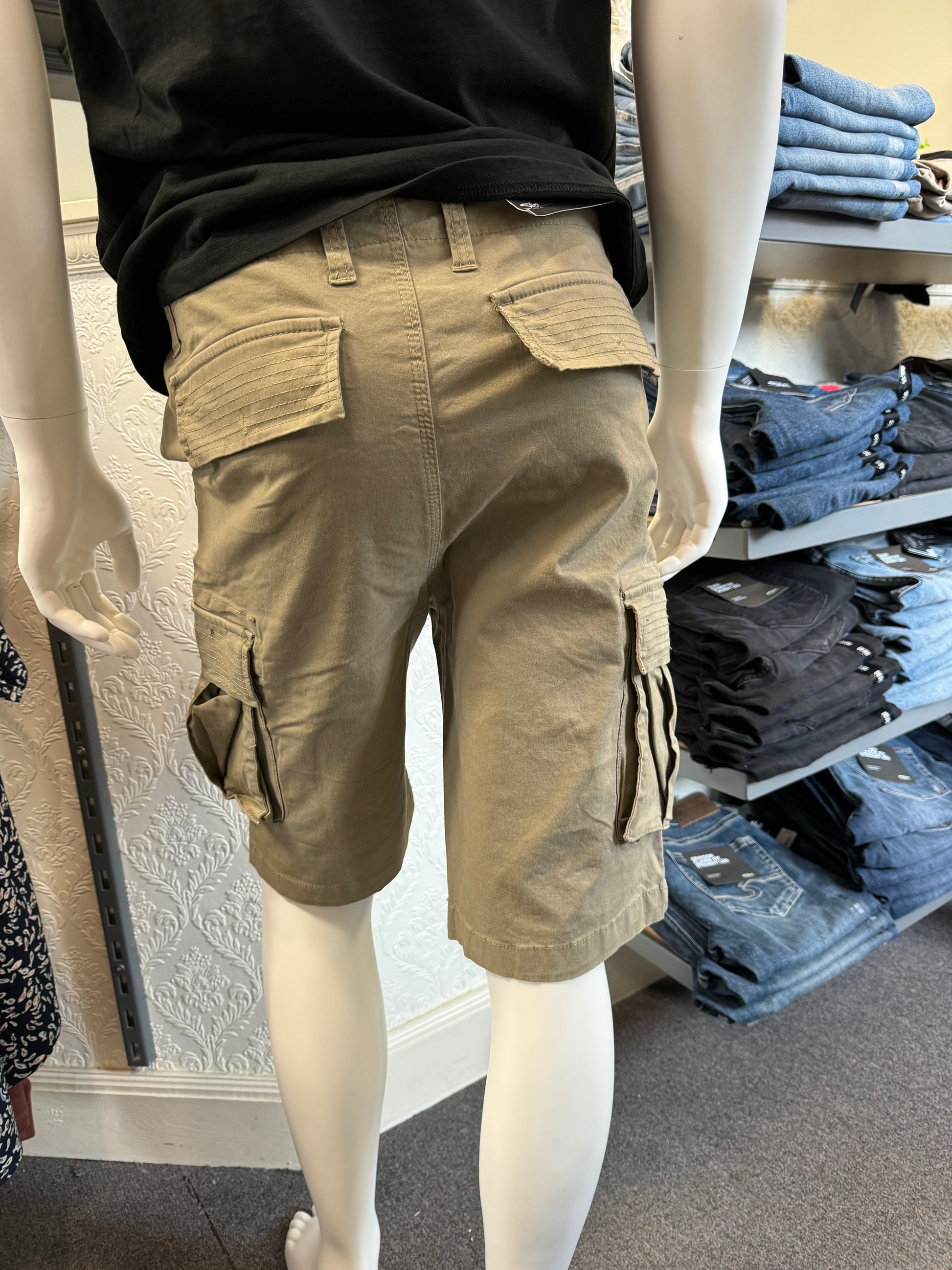 Close-up of the Silver Jeans Collin Classic Fit Cargo Bermudas in Light Olive, distinguished by a large side pocket, displayed on a mannequin. The fabric exhibits detailed stitching and the material looks exceptionally durable. In the background, shelves filled with folded clothes are visible.