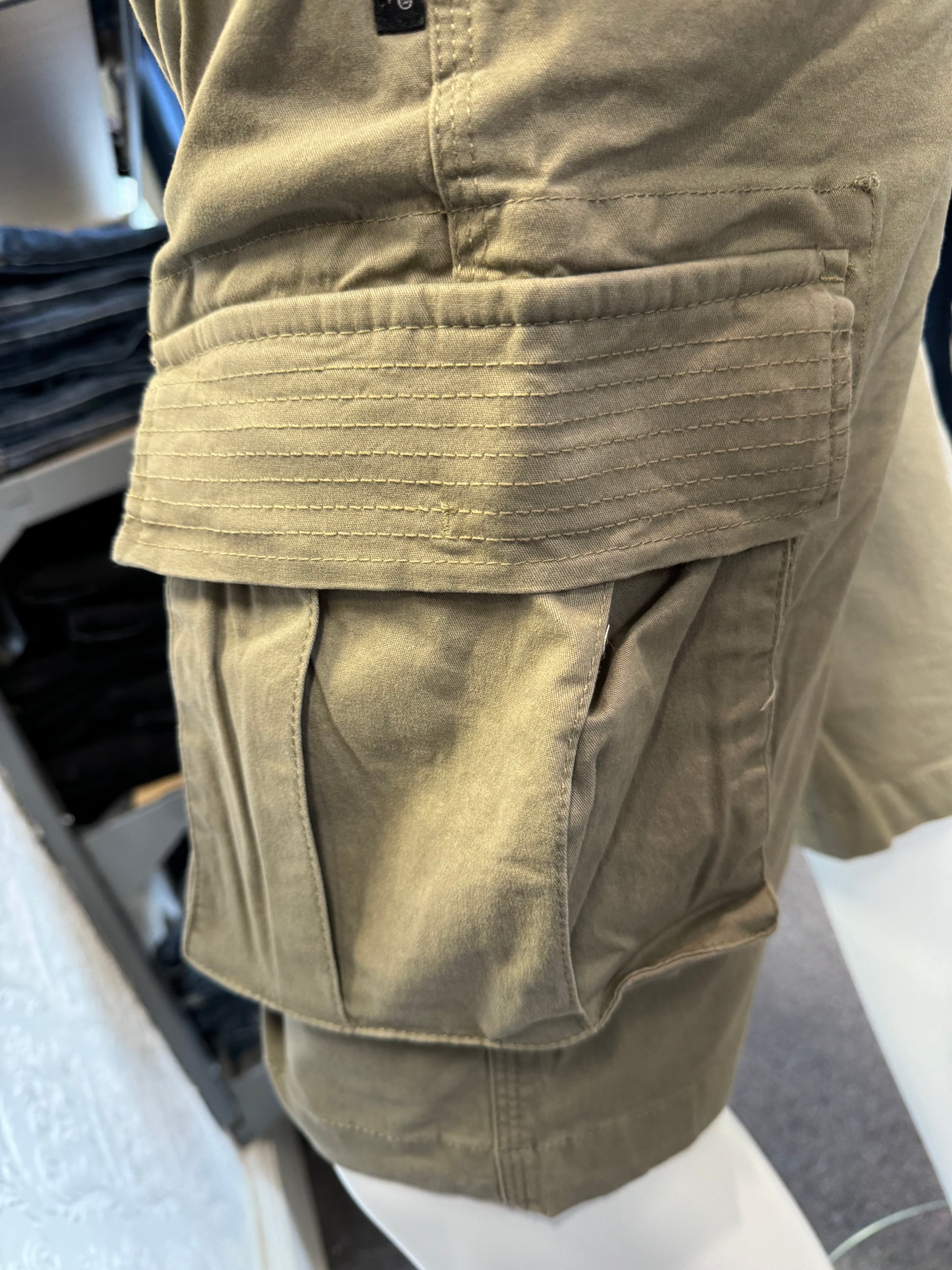 Close-up of the Silver Jeans Collin Classic Fit Cargo Bermudas in Light Olive, distinguished by a large side pocket, displayed on a mannequin. The fabric exhibits detailed stitching and the material looks exceptionally durable. In the background, shelves filled with folded clothes are visible.