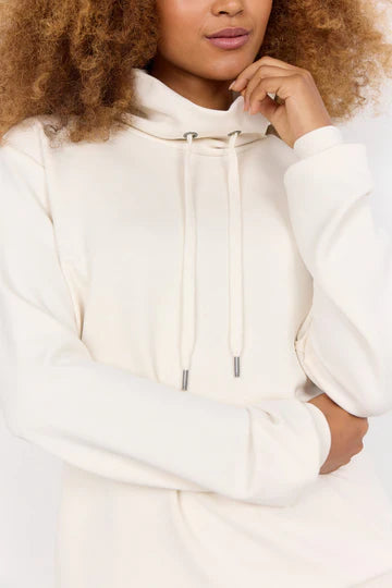 A person with curly hair wears the SC-BANU 125 SWEATSHIRT in cream by SoyaConcept, a light-colored, long-sleeve hoodie featuring a high neckline and drawstrings. Their arm is partially crossed over their chest, exuding a comfortable sweatshirt vibe. The photo is cropped just above their mouth and mid-torso, highlighting the homewear look of the upper body details.