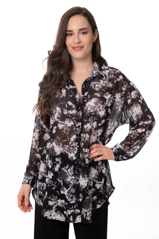 A fashion-forward individual with long, dark, wavy hair poses confidently in a Bali Corp White and Black Flower Print Shirt against a white background. They smile slightly with one hand on their hip, exuding a relaxed and assured demeanor.