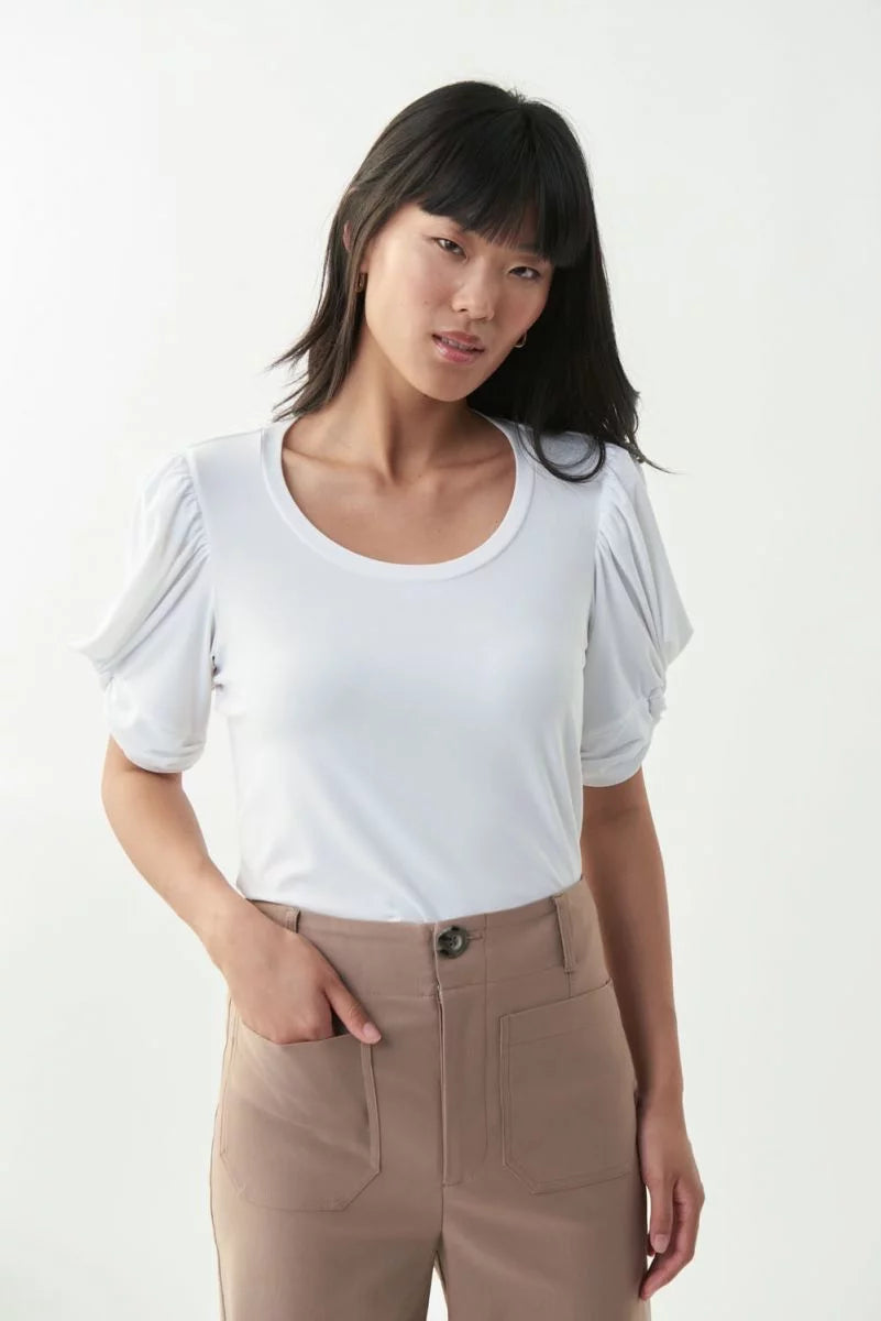 A person with long dark hair is wearing the Elegant White Shirt 221157 by Joseph Ribkoff and beige high-waisted pants. They have their right hand in a pants pocket and are standing against a light-colored background, looking slightly to their left. This must-have outfit exudes effortless style.