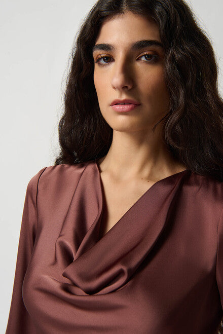 A woman stands against a plain white background. She has long, wavy hair and is wearing Joseph Ribkoff's Draped Satin Blouse Style 234082 in maroon, which features three-quarter sleeves and is crafted from a lightweight satin blend. She pairs it with patterned black and brown leggings and black ankle boots. Her arms are relaxed at her sides.