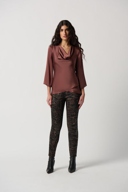 A woman stands against a plain white background. She has long, wavy hair and is wearing Joseph Ribkoff's Draped Satin Blouse Style 234082 in maroon, which features three-quarter sleeves and is crafted from a lightweight satin blend. She pairs it with patterned black and brown leggings and black ankle boots. Her arms are relaxed at her sides.