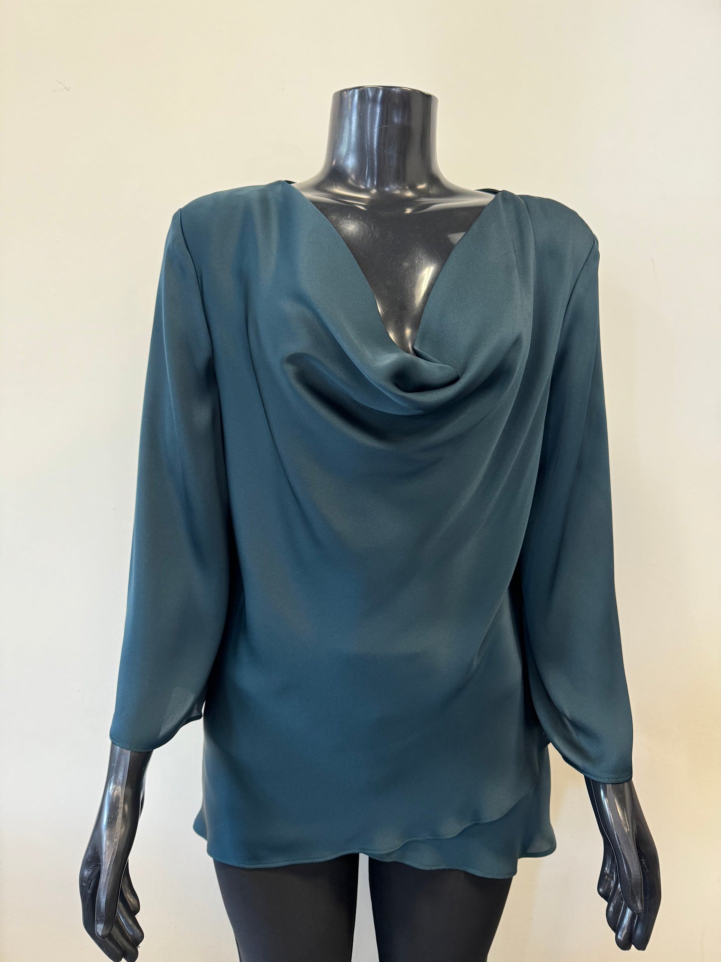 A mannequin dressed in a deep teal, long-sleeve Draped Satin Blouse Style 234082 by Joseph Ribkoff in Alpine Green. The lightweight satin blend blouse features elegant folds and a tiered hem with a draped cowl neckline. The mannequin's black, glossy surface contrasts with the rich color of the blouse. The background is plain.