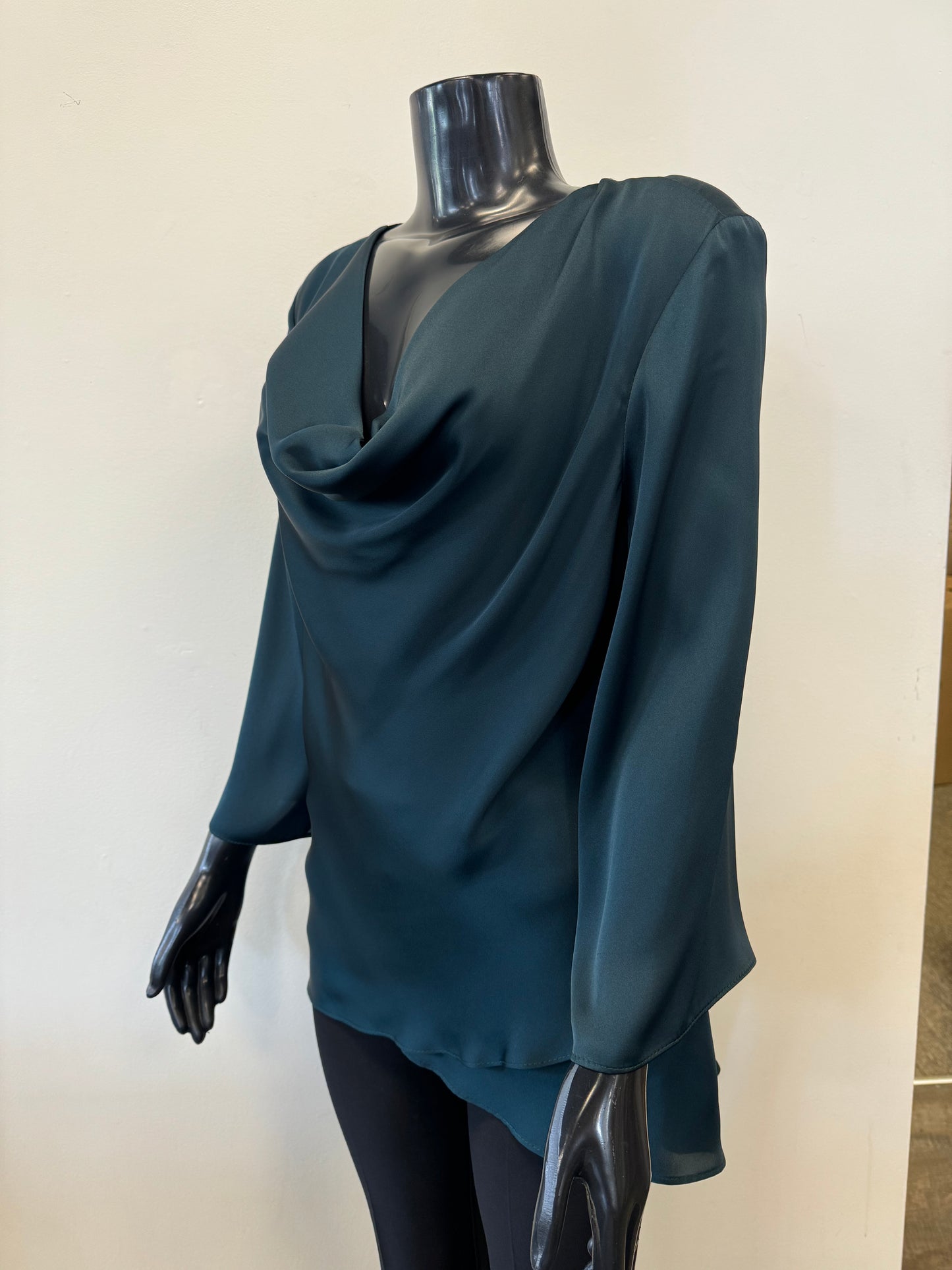 A mannequin dressed in a deep teal, long-sleeve Draped Satin Blouse Style 234082 by Joseph Ribkoff in Alpine Green. The lightweight satin blend blouse features elegant folds and a tiered hem with a draped cowl neckline. The mannequin's black, glossy surface contrasts with the rich color of the blouse. The background is plain.