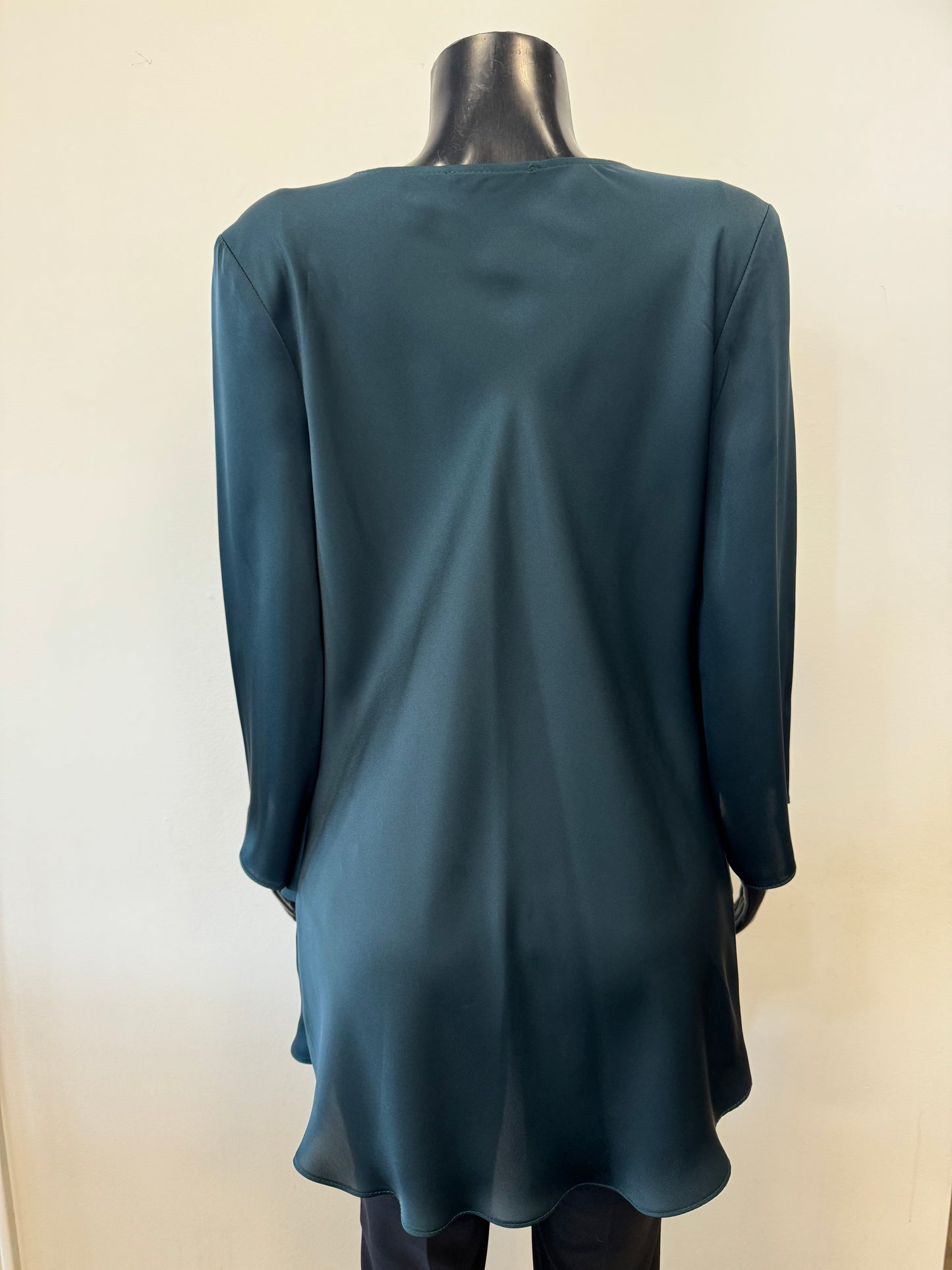 A mannequin dressed in a deep teal, long-sleeve Draped Satin Blouse Style 234082 by Joseph Ribkoff in Alpine Green. The lightweight satin blend blouse features elegant folds and a tiered hem with a draped cowl neckline. The mannequin's black, glossy surface contrasts with the rich color of the blouse. The background is plain.