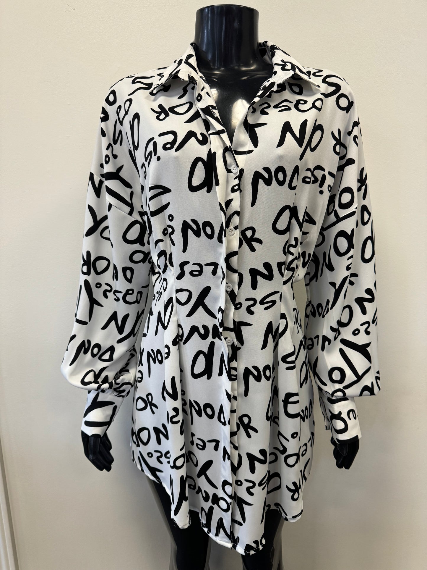 Close-up of a Black And White Word Printed Shirt by ETERNELLE with abstract letters and numbers. The slightly wrinkled fabric features a seam running vertically down the center, showcasing its versatile style. The background includes part of an office environment with a wooden surface and walls, enhancing its sleek design.