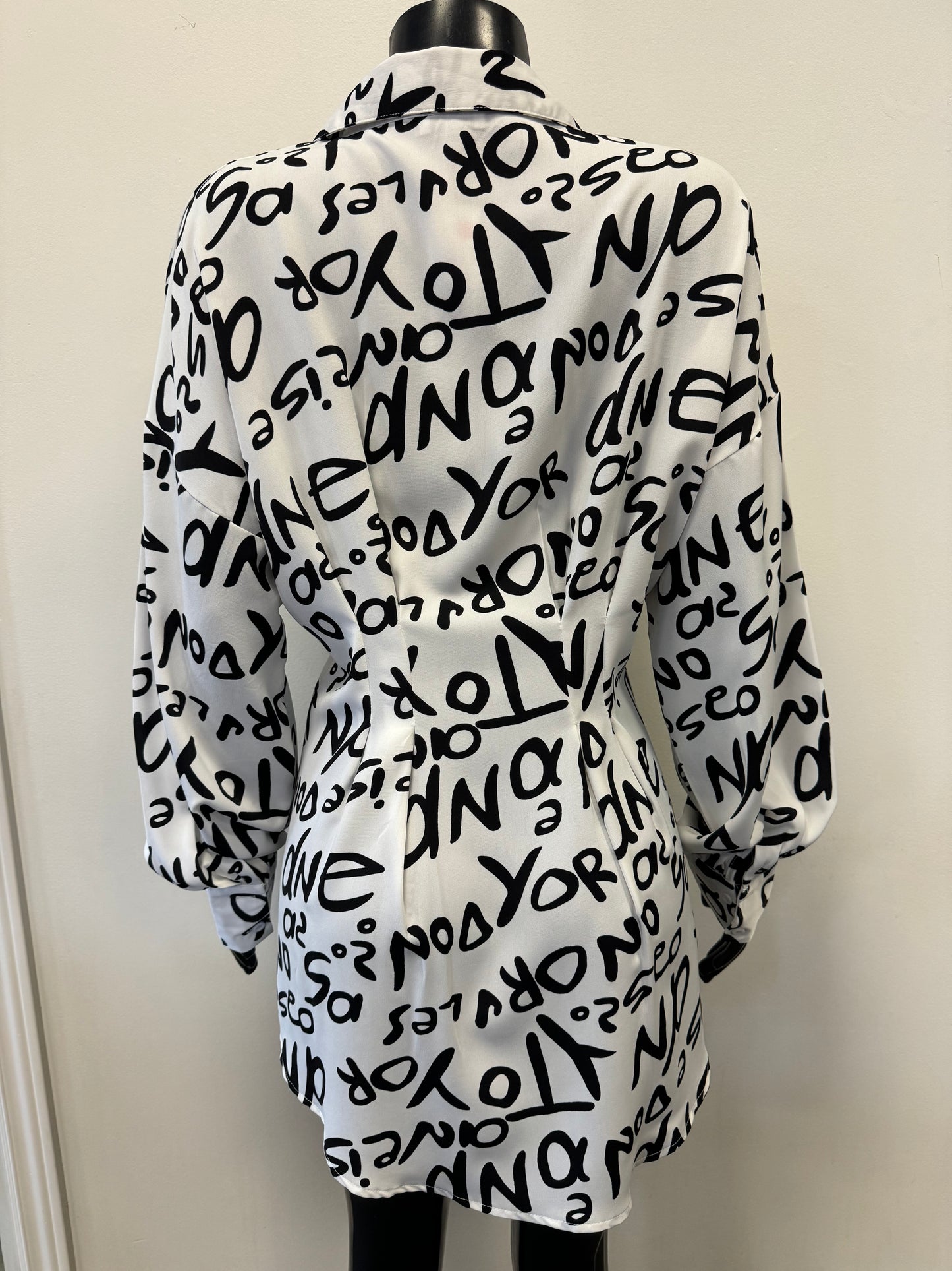 Close-up of a Black And White Word Printed Shirt by ETERNELLE with abstract letters and numbers. The slightly wrinkled fabric features a seam running vertically down the center, showcasing its versatile style. The background includes part of an office environment with a wooden surface and walls, enhancing its sleek design.