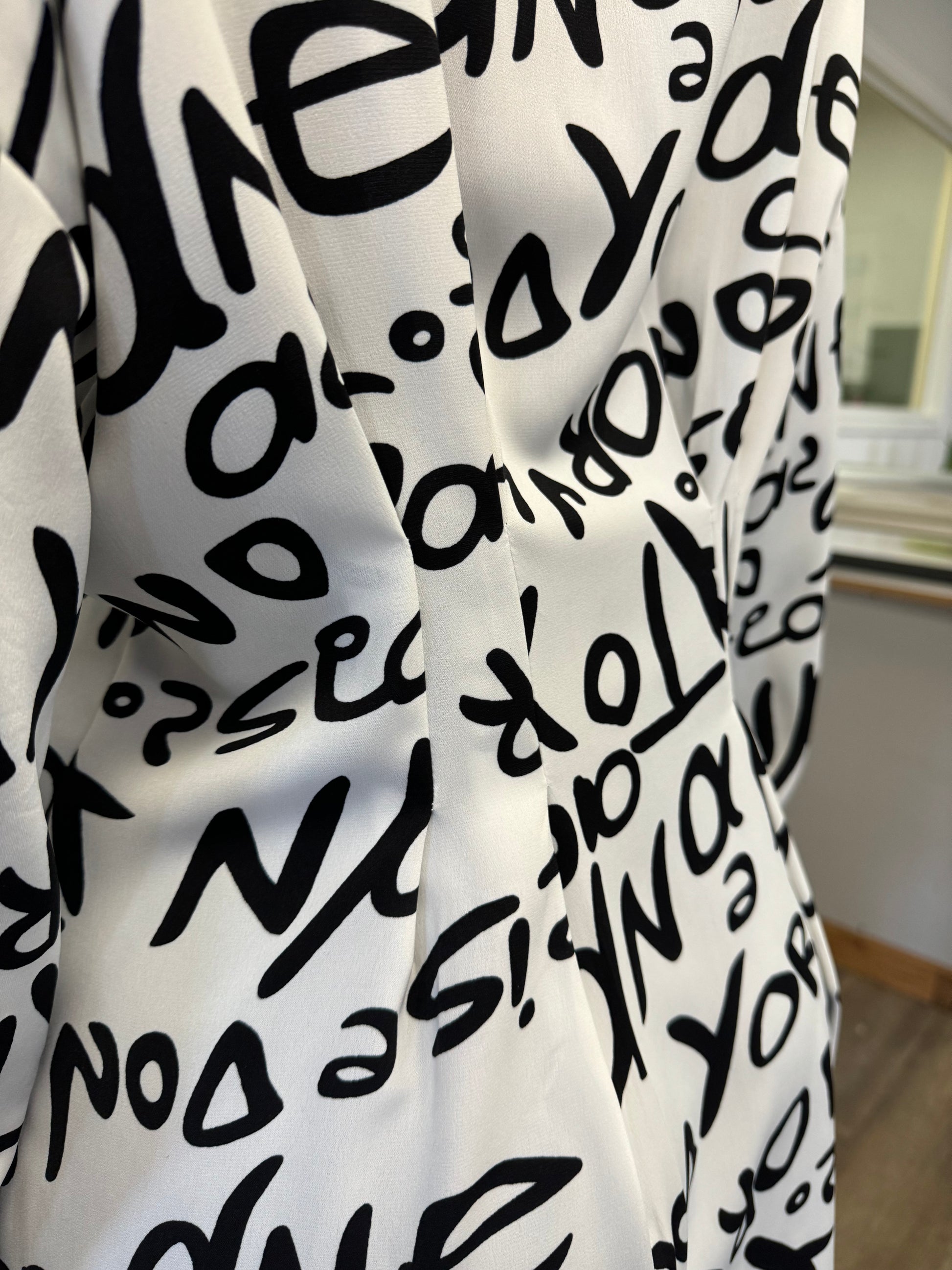 Close-up of a Black And White Word Printed Shirt by ETERNELLE with abstract letters and numbers. The slightly wrinkled fabric features a seam running vertically down the center, showcasing its versatile style. The background includes part of an office environment with a wooden surface and walls, enhancing its sleek design.