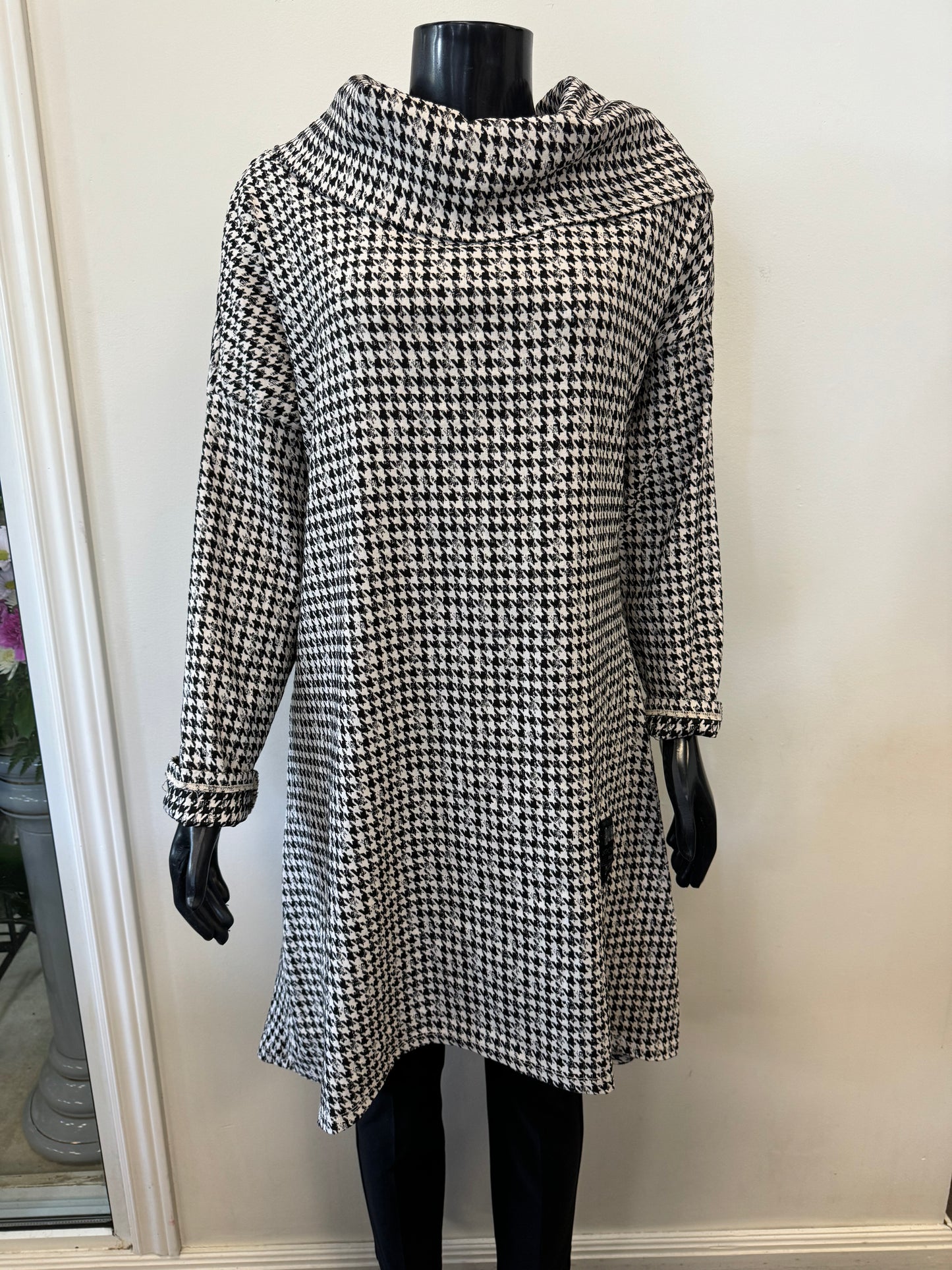 A mannequin displayed in an ETERNELLE Dogtooth Winter Dress, featuring a long-sleeve, knee-length design with a high neckline. The backdrop is a plain white wall, complemented by a glimpse of a potted plant on the left side. This chic piece is crafted in Italy and exudes timeless elegance.