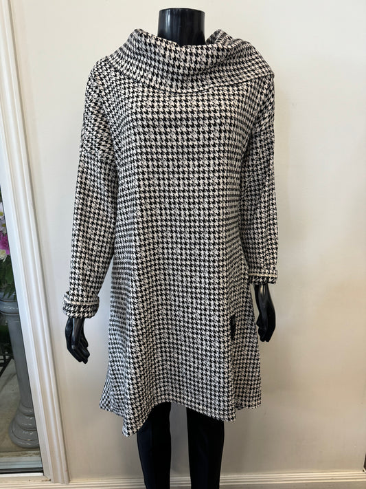 A mannequin displayed in an ETERNELLE Dogtooth Winter Dress, featuring a long-sleeve, knee-length design with a high neckline. The backdrop is a plain white wall, complemented by a glimpse of a potted plant on the left side. This chic piece is crafted in Italy and exudes timeless elegance.