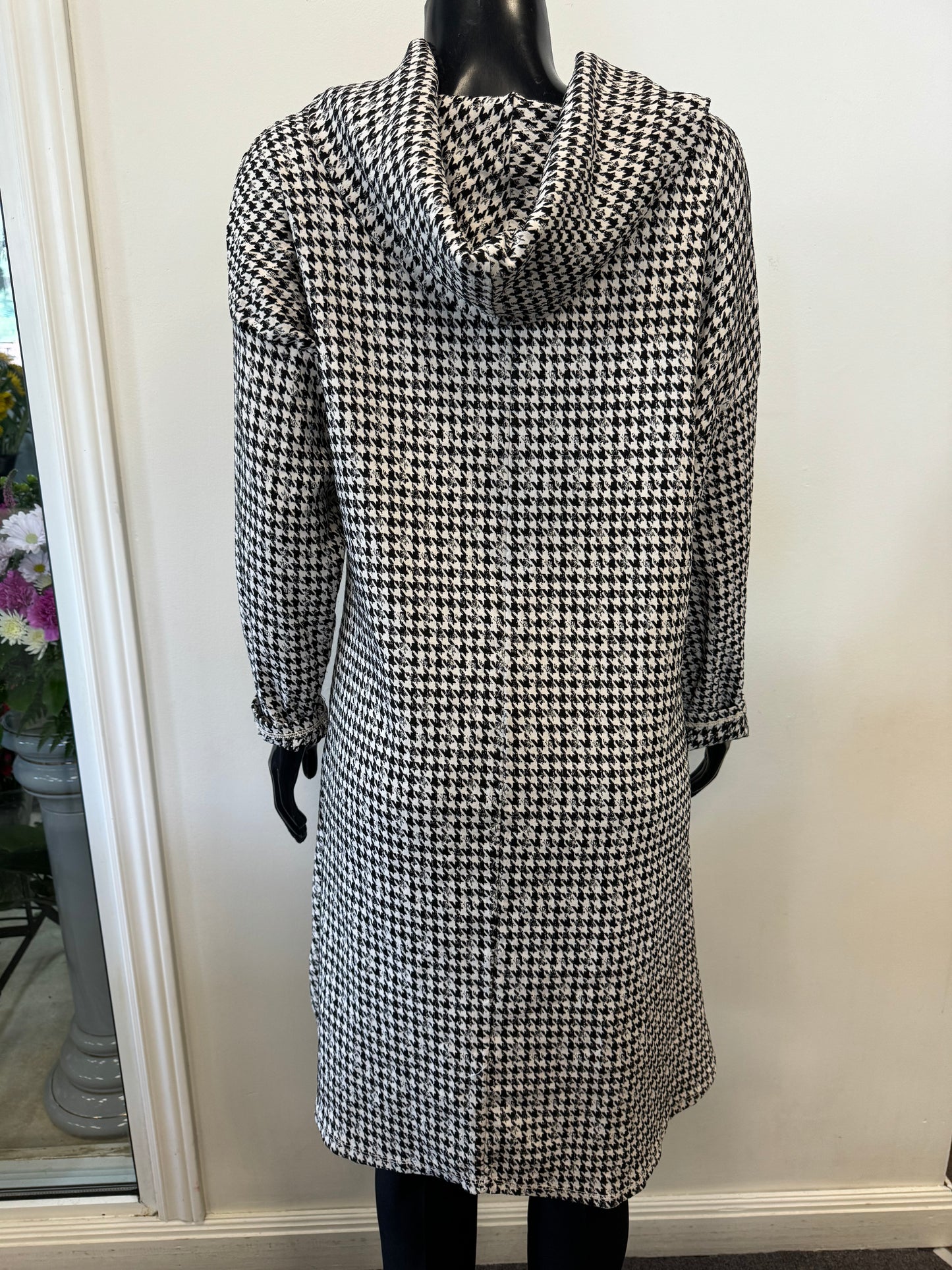 A mannequin displayed in an ETERNELLE Dogtooth Winter Dress, featuring a long-sleeve, knee-length design with a high neckline. The backdrop is a plain white wall, complemented by a glimpse of a potted plant on the left side. This chic piece is crafted in Italy and exudes timeless elegance.