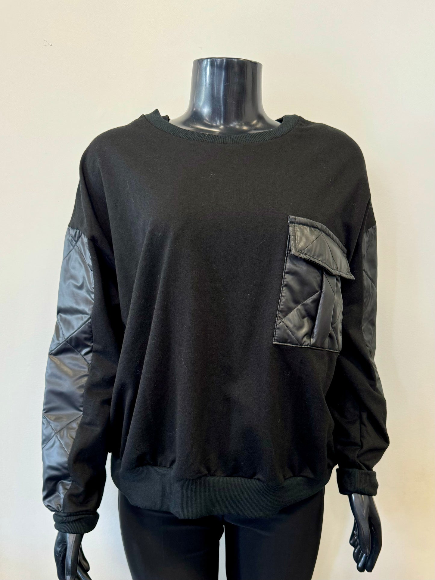 A stylish sweater from ETERNELLE, known as the Sweater with Pocket - ETERNELLE, is elegantly showcased on a black mannequin. The cozy pullover features quilted nylon patches on the sleeves and a large square quilted nylon pocket on the left side of the chest. Its simple, casual design complements the soft and comfortable fabric perfectly.