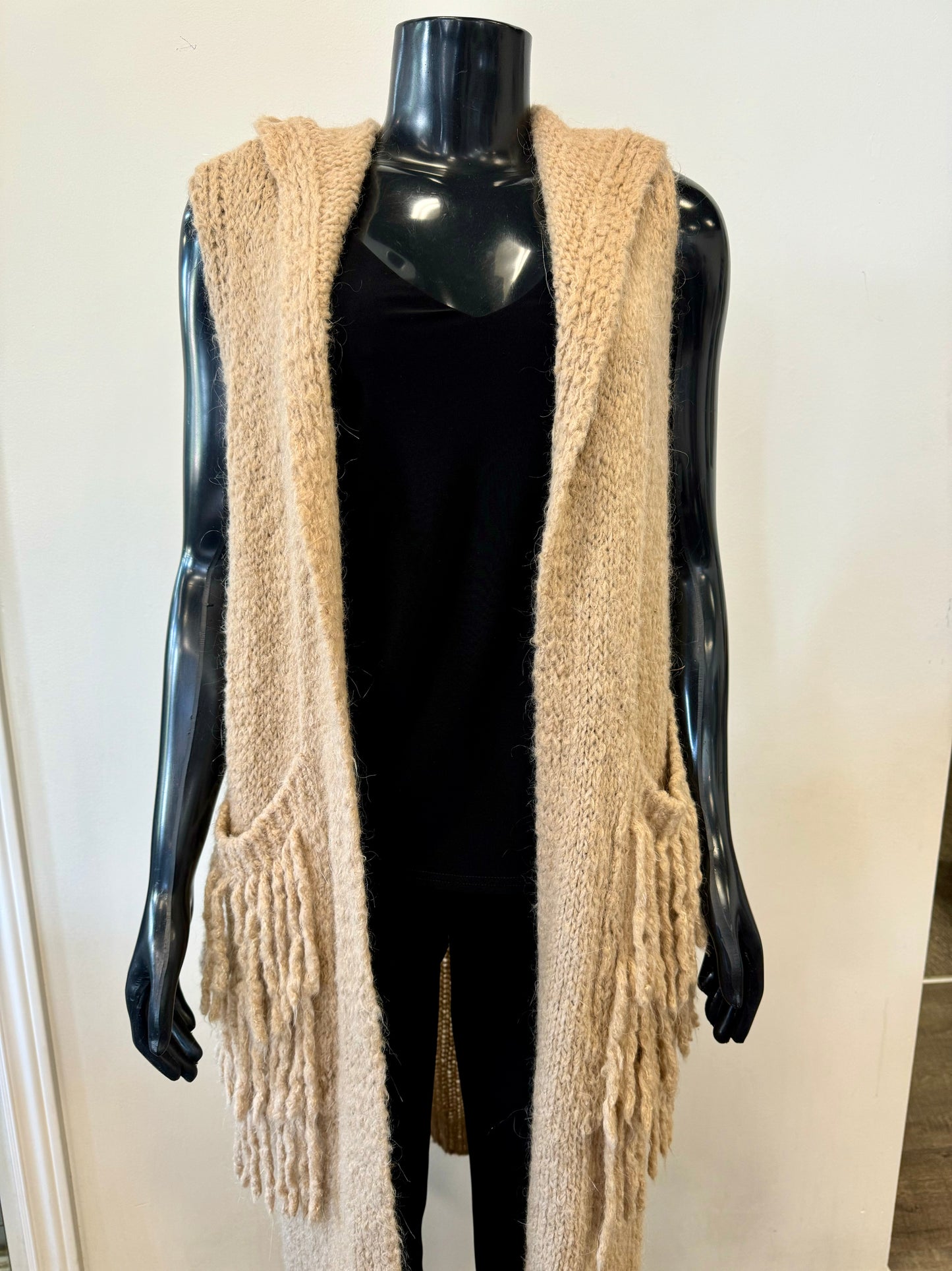 A black mannequin is dressed in the ETERNELLE Large Knit Cardigan, a luxurious blend of beige in a sleeveless design with a hood and a warm open front. The cardigan features two large fringed pockets on either side. Underneath, the mannequin is wearing a black top and black pants against a plain white wall backdrop.