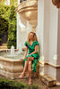 A woman with curly blonde hair, wearing a green Joseph Ribkoff Floral Print Silky Knit Flowy Wrap Dress with ruffled sleeves, sits barefoot on the edge of a stone fountain in a sunlit courtyard. She gazes thoughtfully at the water beside her. Lush greenery and architectural details fill the background.