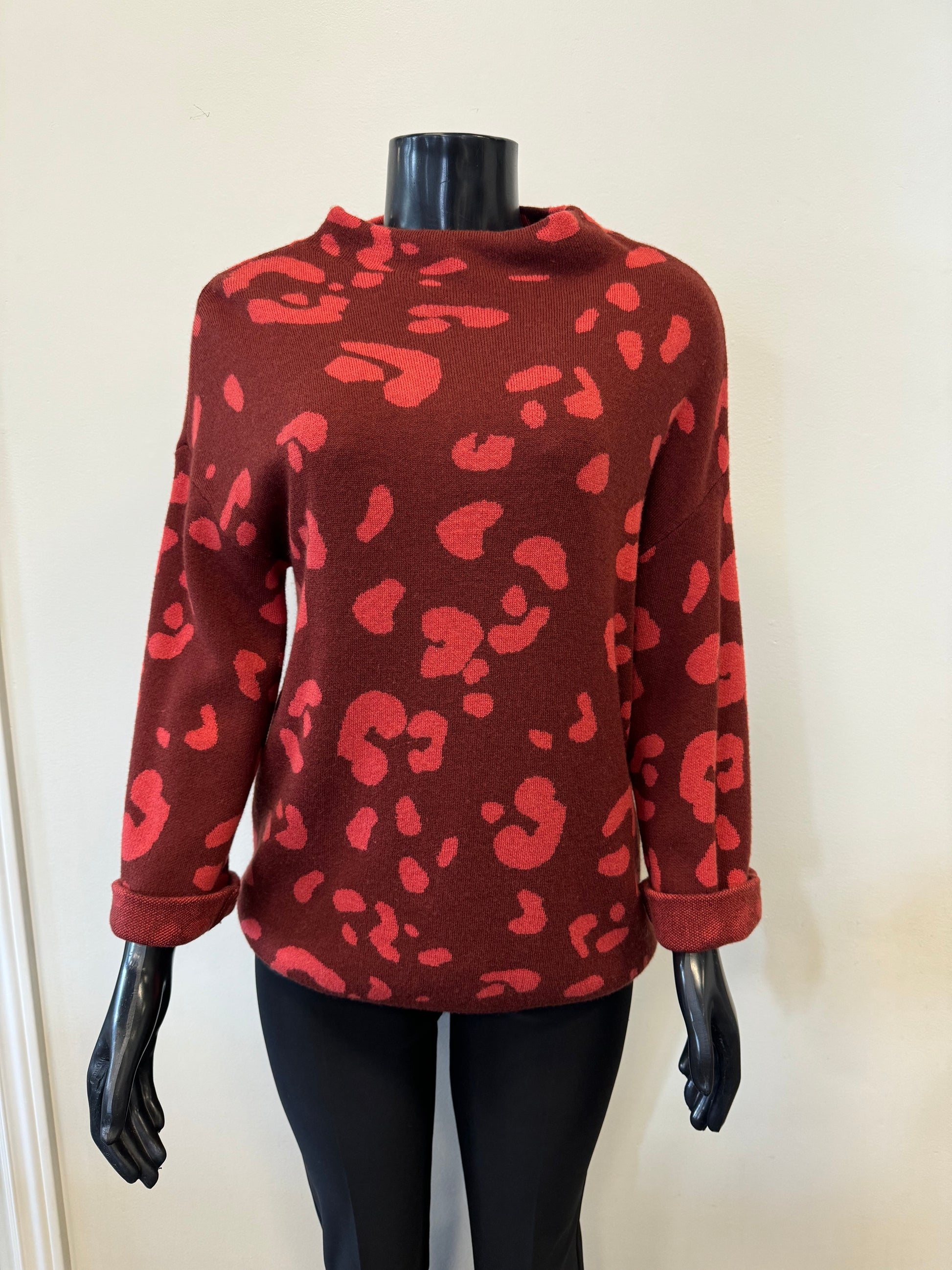 A mannequin sports a luxurious ensemble featuring the Tribal Knit Sweater Patterned - Tribal GARNENT in vibrant red with darker abstract leopard prints. The long sleeves pair effortlessly with sleek black leggings against a plain light-colored wall, radiating an effortlessly chic vibe.