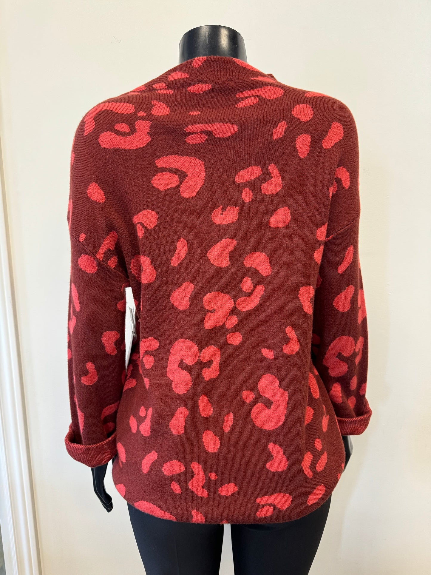 A mannequin stands against a plain wall wearing the Snit Sweater Patterned - Tribal GARNENT from the brand Tribal. This luxurious blend sweater features a bold, abstract pattern of large, irregular darker red spots and has a relaxed fit with rolled-up sleeves. The outfit is completed with black pants, embodying both comfort and style.