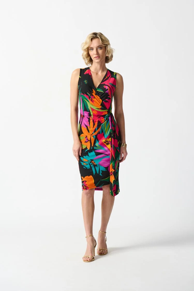 A blonde woman is wearing Joseph Ribkoff's Silky Knit Tropical Print Wrap Dress (style 242012 Hots/Hero) which boasts vibrant hues of orange, pink, and green. The dress features a sleeveless design, a tie at the waist, and a ruffled skirt. She is standing on a plain white background and paired it with nude high heel sandals.