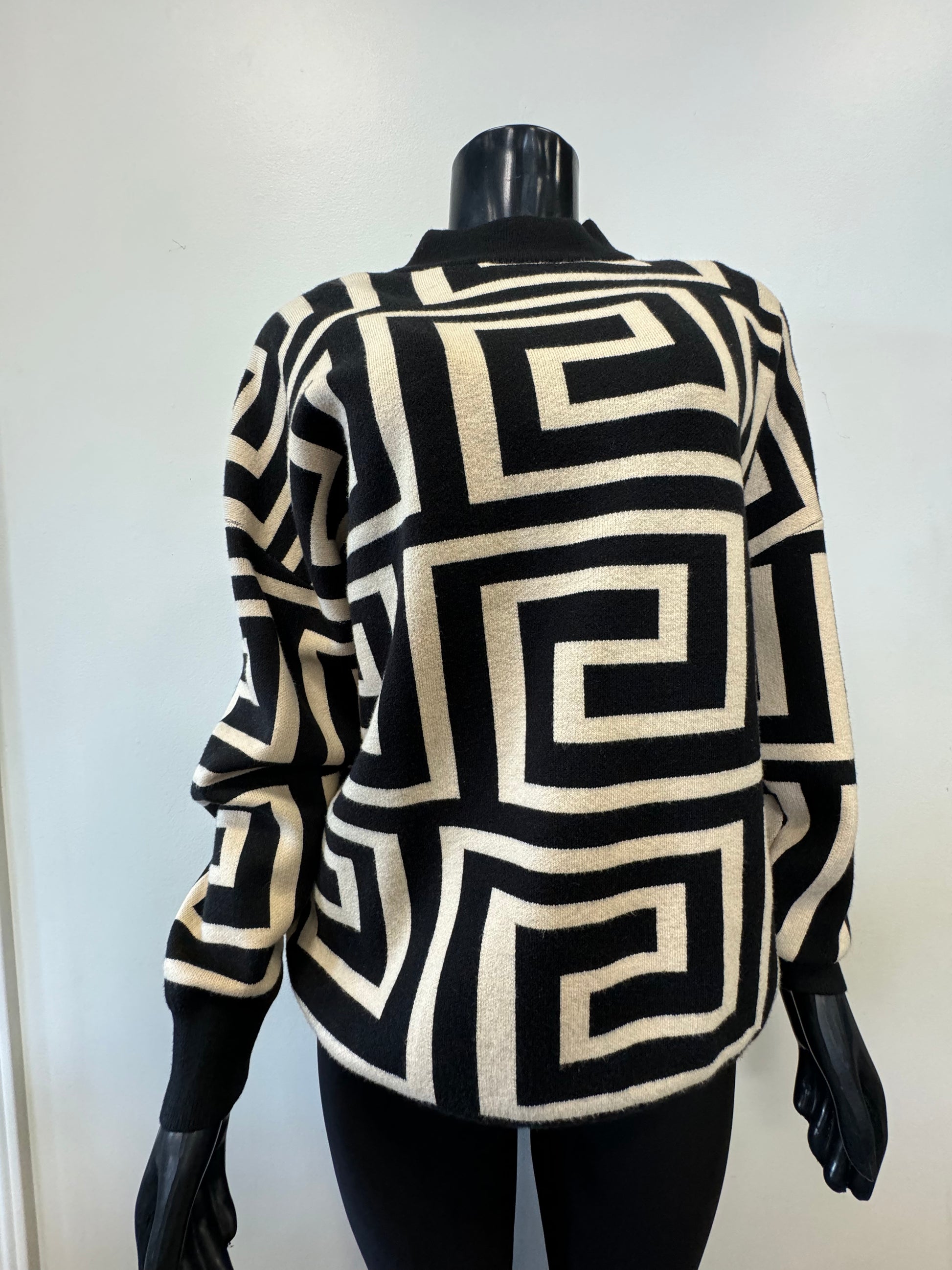 A black mannequin is dressed in the Knit Sweater Block Pattern by ETERNELLE, showcasing a striking geometric block design with black and white square spirals. The sweater features a high neckline, and the background wall behind the mannequin is plain and light-colored.