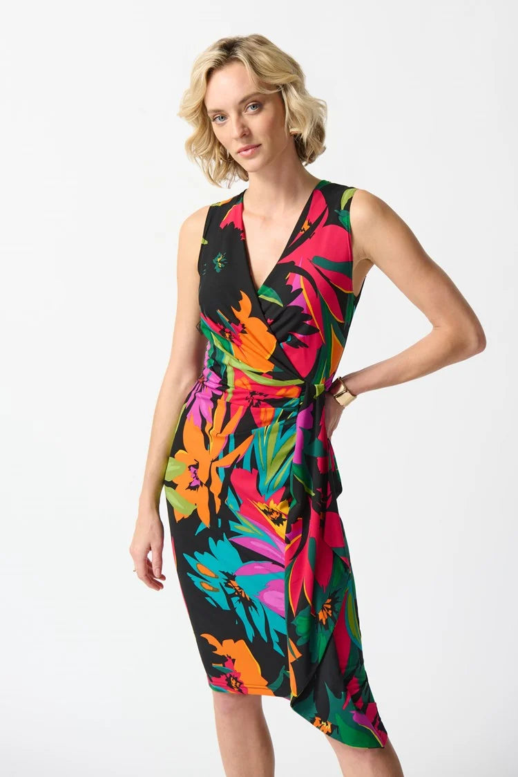 A blonde woman is wearing Joseph Ribkoff's Silky Knit Tropical Print Wrap Dress (style 242012 Hots/Hero) which boasts vibrant hues of orange, pink, and green. The dress features a sleeveless design, a tie at the waist, and a ruffled skirt. She is standing on a plain white background and paired it with nude high heel sandals.