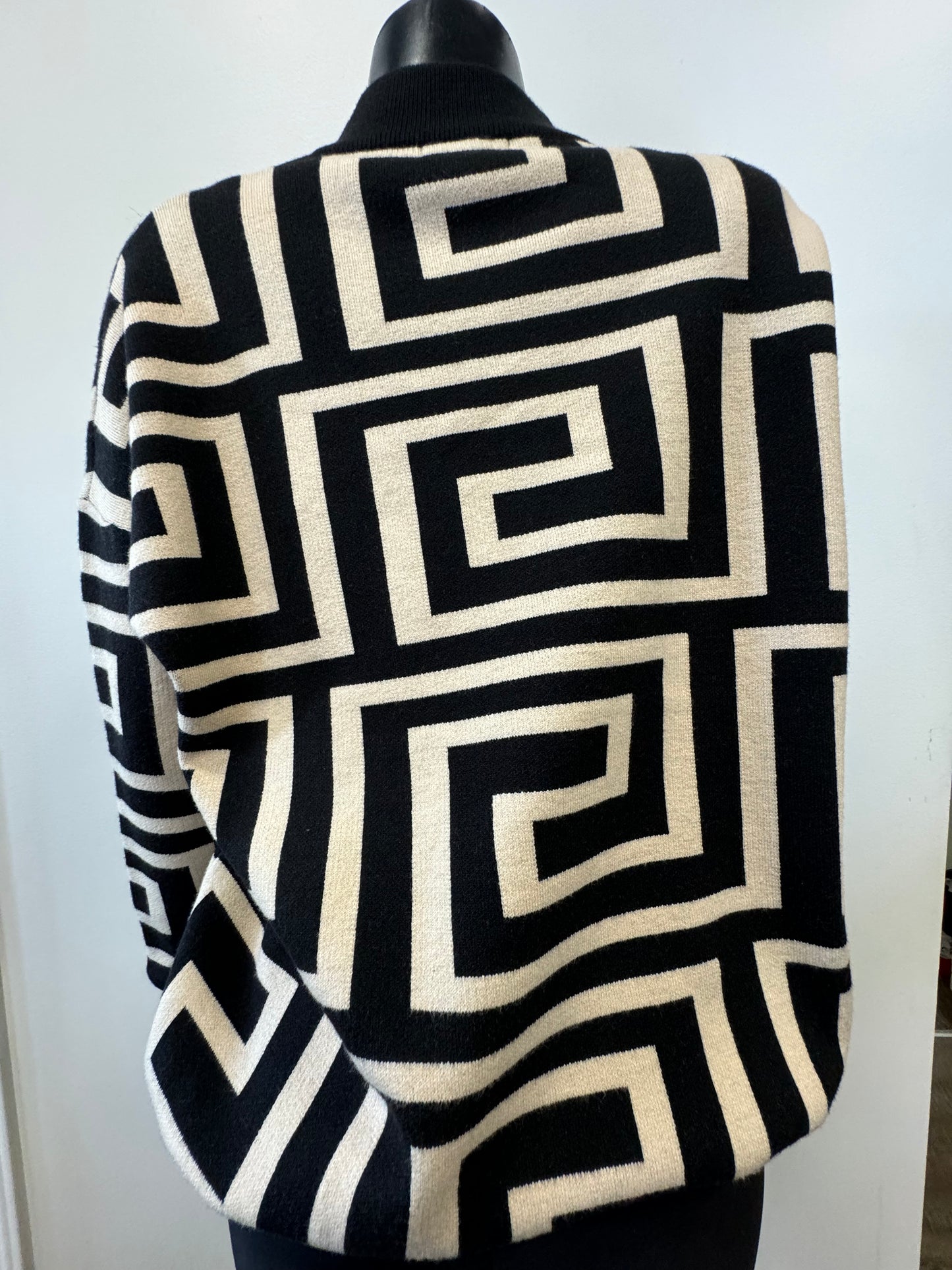 A black mannequin is dressed in the Knit Sweater Block Pattern by ETERNELLE, showcasing a striking geometric block design with black and white square spirals. The sweater features a high neckline, and the background wall behind the mannequin is plain and light-colored.