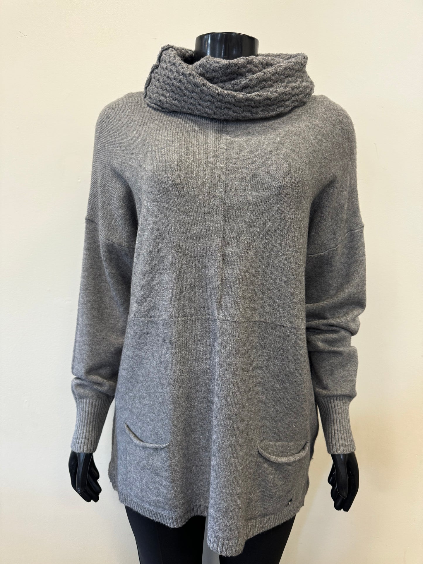 The "Knit Sweater with Scarf - ETERNELLE" by ETERNELLE, featuring a gray knit design and a thick, ribbed cowl neck resembling a cozy scarf, is shown on a black mannequin. The lightweight fabric includes long sleeves, a relaxed fit, and two small front pockets. The background is plain and light-colored.