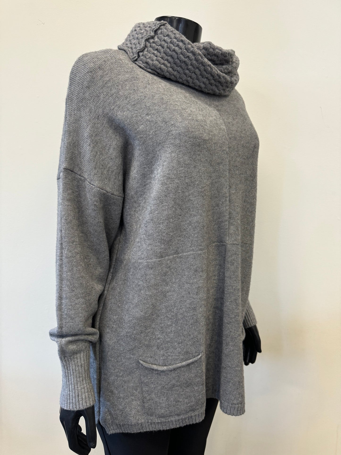 The "Knit Sweater with Scarf - ETERNELLE" by ETERNELLE, featuring a gray knit design and a thick, ribbed cowl neck resembling a cozy scarf, is shown on a black mannequin. The lightweight fabric includes long sleeves, a relaxed fit, and two small front pockets. The background is plain and light-colored.