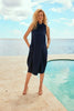 A woman with curly blonde hair stands by a pool overlooking the ocean and a partly cloudy sky. She is wearing the Joseph Ribkoff Textured Woven Sleeveless Cocoon Dress 241204 Hots/Magazine Style, paired with beige high-heeled sandals. Her hands are relaxed, and her expression is calm as she gazes at the camera.