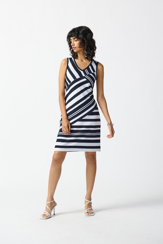 A woman is wearing the Joseph Ribkoff Jacquard Knit Sleeveless Sheath Dress (242077), which features a knee-length shift silhouette with a navy blue and white striped pattern. She stands against a plain white background, accessorized with white strappy high-heeled sandals and a bracelet on her right wrist, looking to her left.