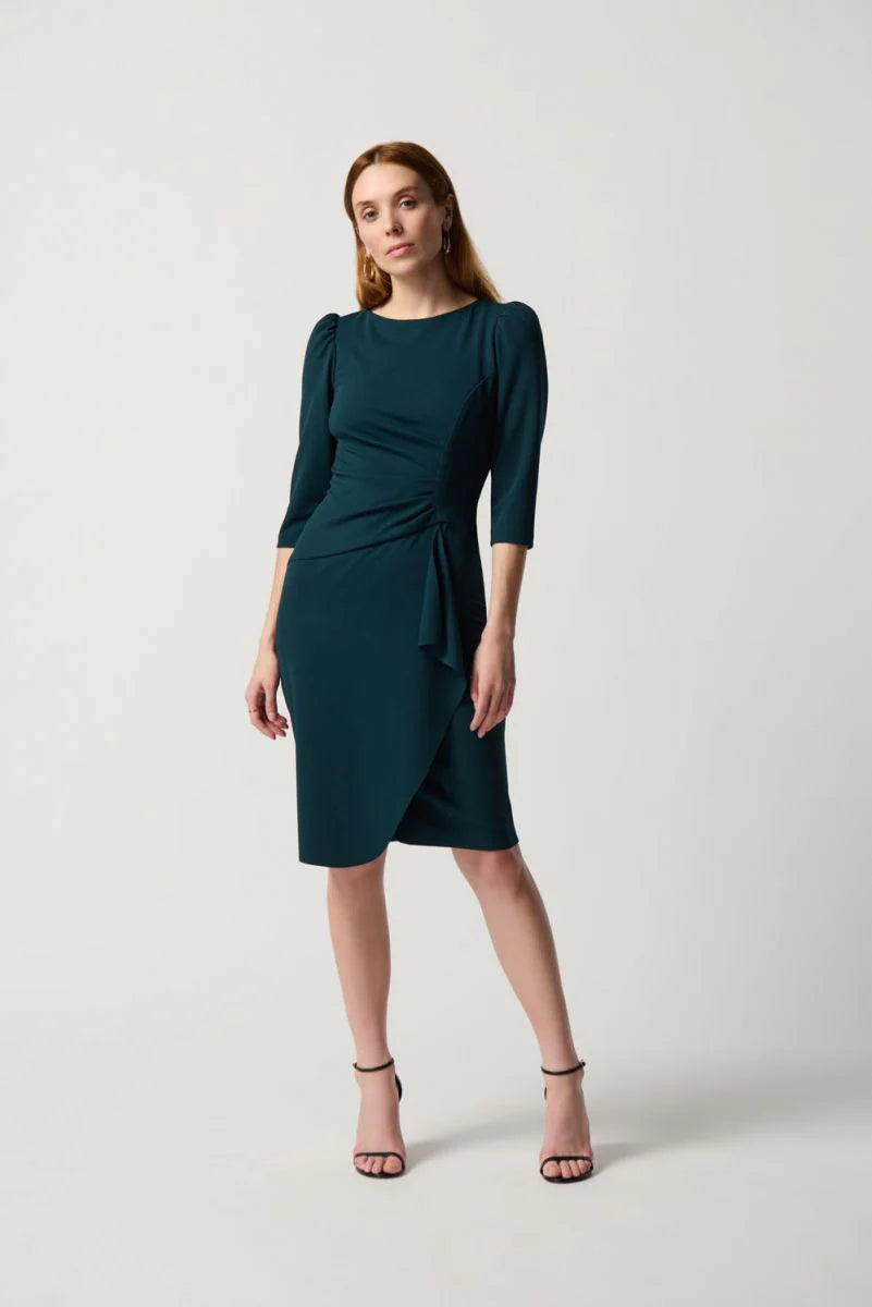 A woman stands against a plain white background, wearing the Boat Neck Dress Style 234290 by Joseph Ribkoff, which is dark green and midi-length with three-quarter sleeves, a boat neck, and a gathered waist detail. She pairs the dress with black high-heeled sandals. She has long, straight, light brown hair and a neutral expression.