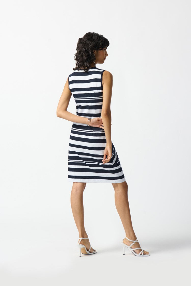 A woman is wearing the Joseph Ribkoff Jacquard Knit Sleeveless Sheath Dress (242077), which features a knee-length shift silhouette with a navy blue and white striped pattern. She stands against a plain white background, accessorized with white strappy high-heeled sandals and a bracelet on her right wrist, looking to her left.