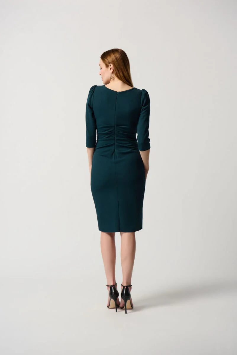 A woman stands against a plain white background, wearing the Boat Neck Dress Style 234290 by Joseph Ribkoff, which is dark green and midi-length with three-quarter sleeves, a boat neck, and a gathered waist detail. She pairs the dress with black high-heeled sandals. She has long, straight, light brown hair and a neutral expression.