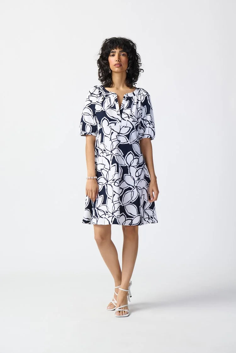 A person with curly hair is posing against a plain background, wearing the Leaf Print Scuba Crepe Fit-and-Flare Dress 241006 by Joseph Ribkoff, adorned with a white and navy blue floral pattern. They have paired it with white peep-toe heels. The dress features quarter-length sleeves and a V-neckline.