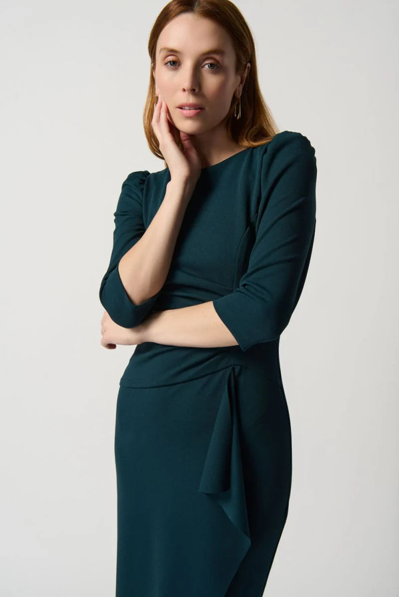 A woman stands against a plain white background, wearing the Boat Neck Dress Style 234290 by Joseph Ribkoff, which is dark green and midi-length with three-quarter sleeves, a boat neck, and a gathered waist detail. She pairs the dress with black high-heeled sandals. She has long, straight, light brown hair and a neutral expression.