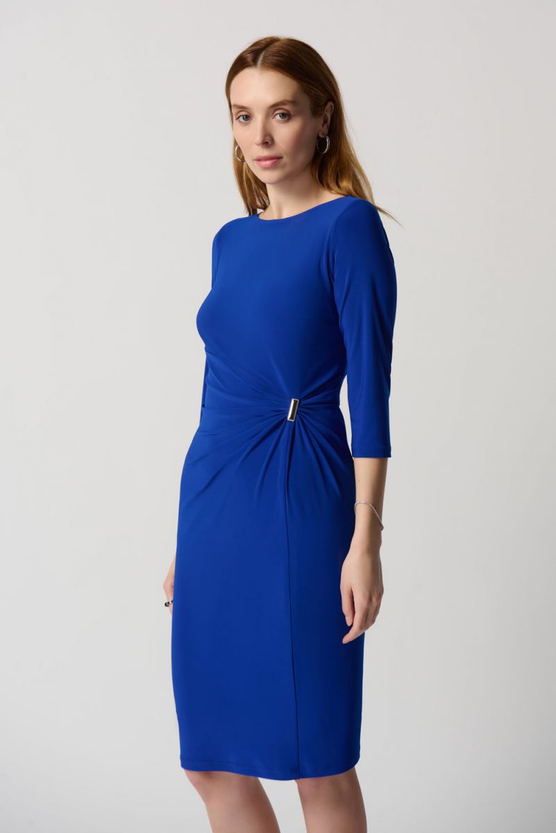 A woman stands against a plain backdrop, wearing a knee-length Gathered Front Dress Style 234031 by Joseph Ribkoff in royal blue with three-quarter sleeves. She has long, straight hair and is wearing open-toe heels. Her hands are gently clasped in front of her, and she has a calm expression on her face.