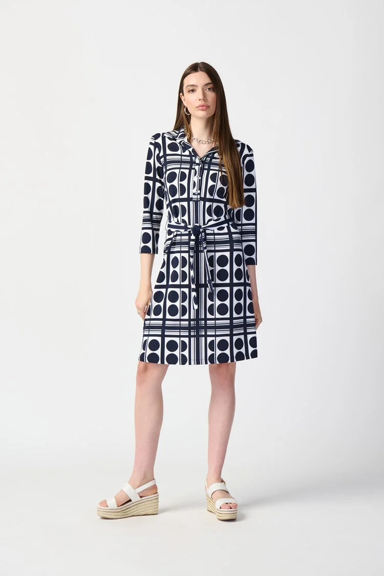 A woman stands wearing the Joseph Ribkoff Dot Print Silky Knit Belted Shirt Dress 241001, which is knee-length with a navy and white geometric pattern. The dress features three-quarter sleeves and includes a matching waist tie that creates a flattering silhouette. She has long, straight brown hair and is paired with white platform sandals. The background is plain white.