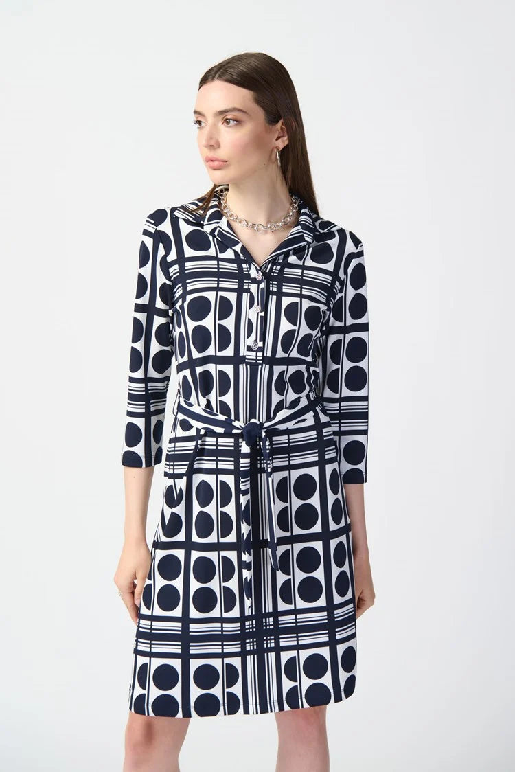 A woman stands wearing the Joseph Ribkoff Dot Print Silky Knit Belted Shirt Dress 241001, which is knee-length with a navy and white geometric pattern. The dress features three-quarter sleeves and includes a matching waist tie that creates a flattering silhouette. She has long, straight brown hair and is paired with white platform sandals. The background is plain white.