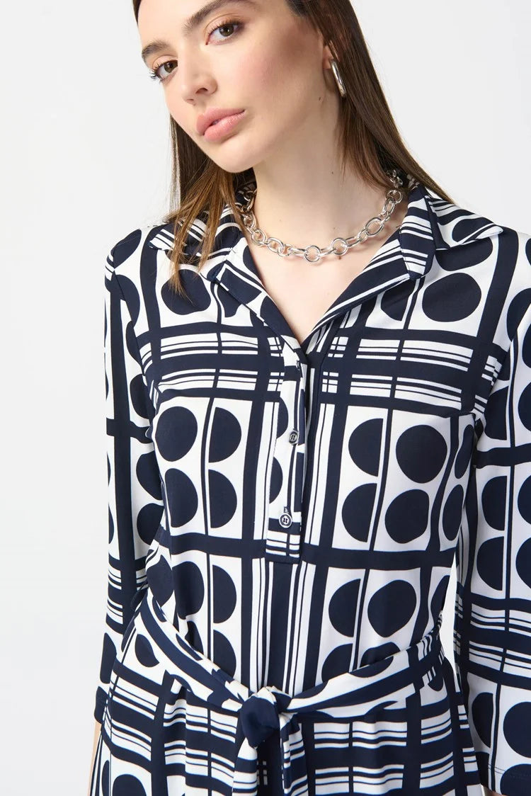 A woman stands wearing the Joseph Ribkoff Dot Print Silky Knit Belted Shirt Dress 241001, which is knee-length with a navy and white geometric pattern. The dress features three-quarter sleeves and includes a matching waist tie that creates a flattering silhouette. She has long, straight brown hair and is paired with white platform sandals. The background is plain white.