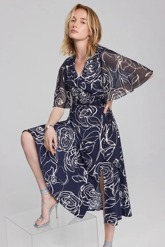 A blonde woman is posing in a Floral Print Silky Knit and Chiffon Dress 241764 from Joseph Ribkoff. The dress, part of the Signature Collection, features short sheer cape sleeves and a knee-length hem. She is seated with one hand on her knee and the other on her hip, wearing light blue strappy high-heeled sandals.