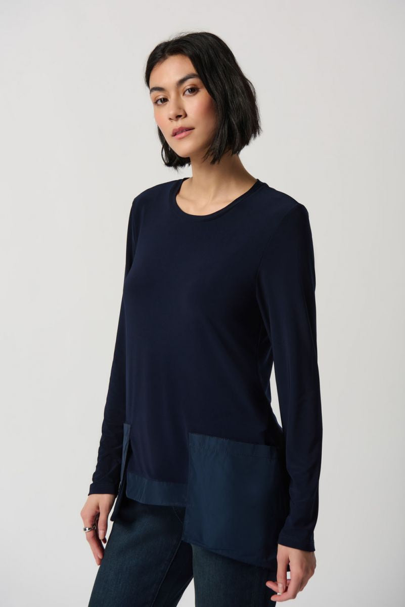 A woman with shoulder-length dark hair wears the Midnight Blue Silky Knit High Low Top Style 234138 by Joseph Ribkoff, featuring a crew neck and asymmetrical hem details. She pairs it with dark jeans and stands against a plain white background, looking towards the camera with a neutral expression. Perfect for winter 2023.