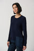 A woman with shoulder-length dark hair wears the Midnight Blue Silky Knit High Low Top Style 234138 by Joseph Ribkoff, featuring a crew neck and asymmetrical hem details. She pairs it with dark jeans and stands against a plain white background, looking towards the camera with a neutral expression. Perfect for winter 2023.