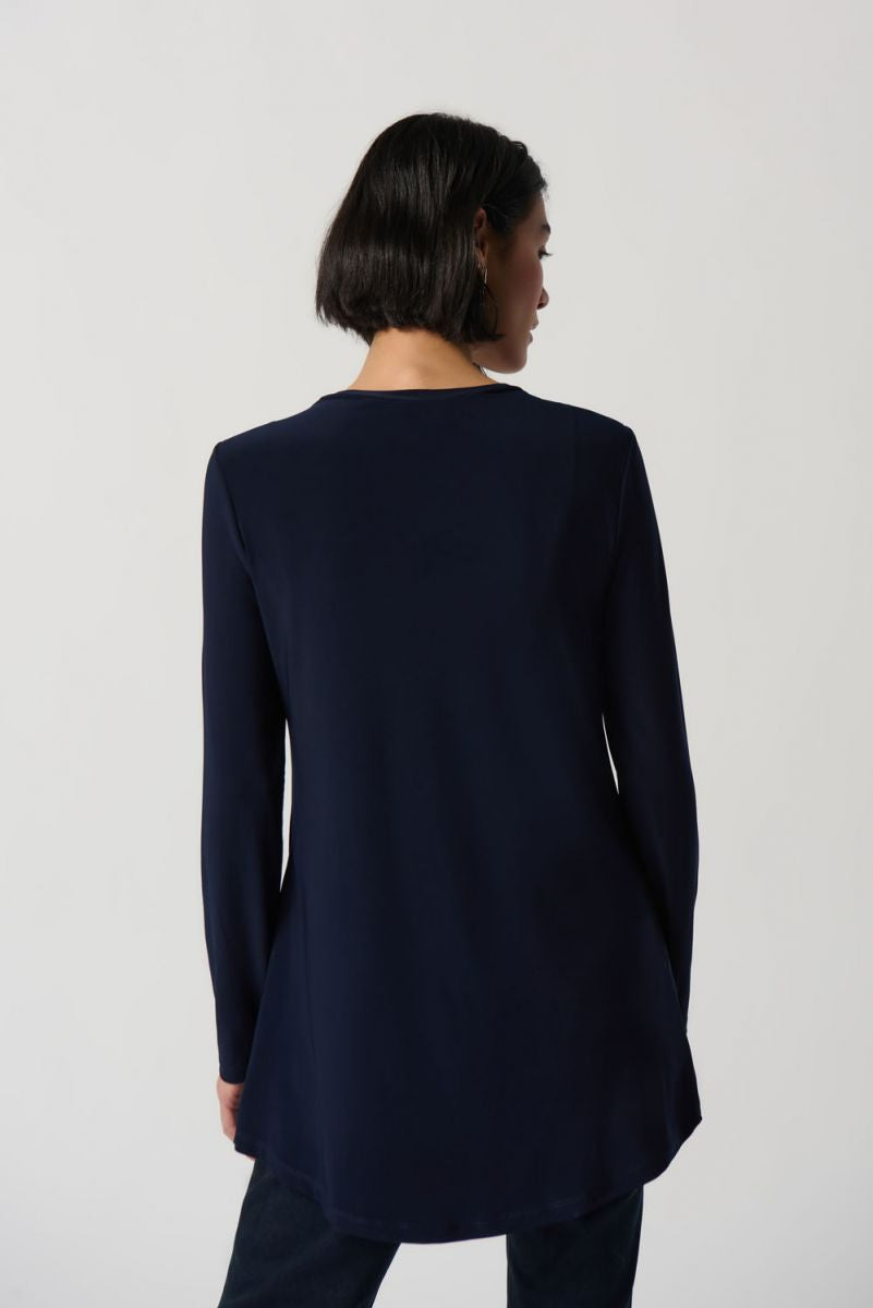 A woman with shoulder-length dark hair wears the Midnight Blue Silky Knit High Low Top Style 234138 by Joseph Ribkoff, featuring a crew neck and asymmetrical hem details. She pairs it with dark jeans and stands against a plain white background, looking towards the camera with a neutral expression. Perfect for winter 2023.