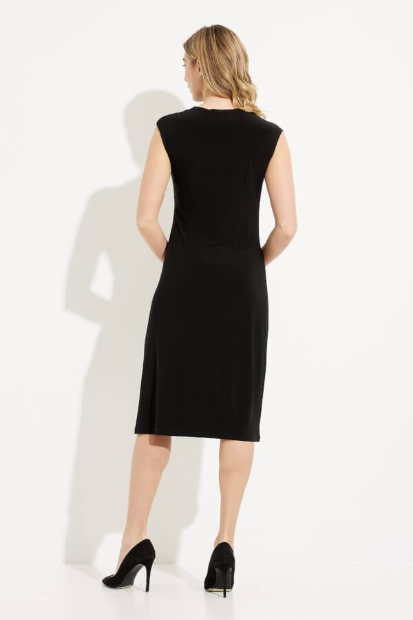 A woman with long, blonde hair stands against a plain white background, wearing the Draped Neck Dress Style 233211 by Joseph Ribkoff. The sleeveless black dress features a draped neckline and a knee-length hem with a small slit on one side. Her left hand touches her chin while her right hand rests on her hip.